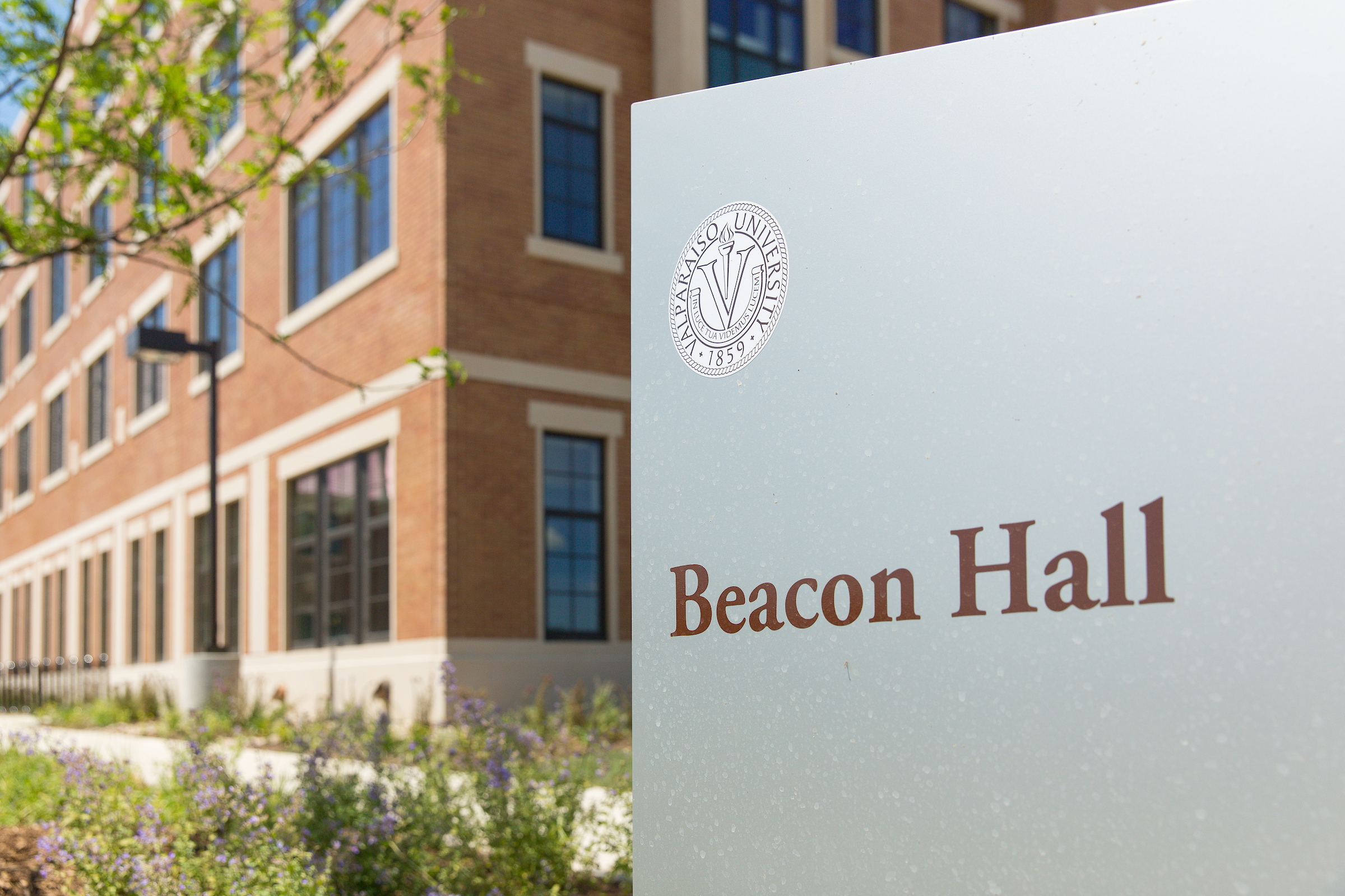 A sign reading "Beacon Hall" stands in front of a brick university building on the Valpo campus, capturing a glimpse of vibrant campus life on a sunny day.