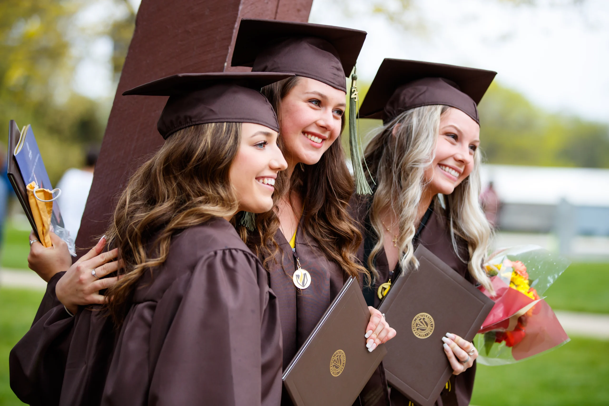 Three smiling graduates in brown caps and gowns pose outdoors, holding diplomas and flowers—capturing a proud moment to share with family, friends, or those seeking Info for Faculty about commencement.