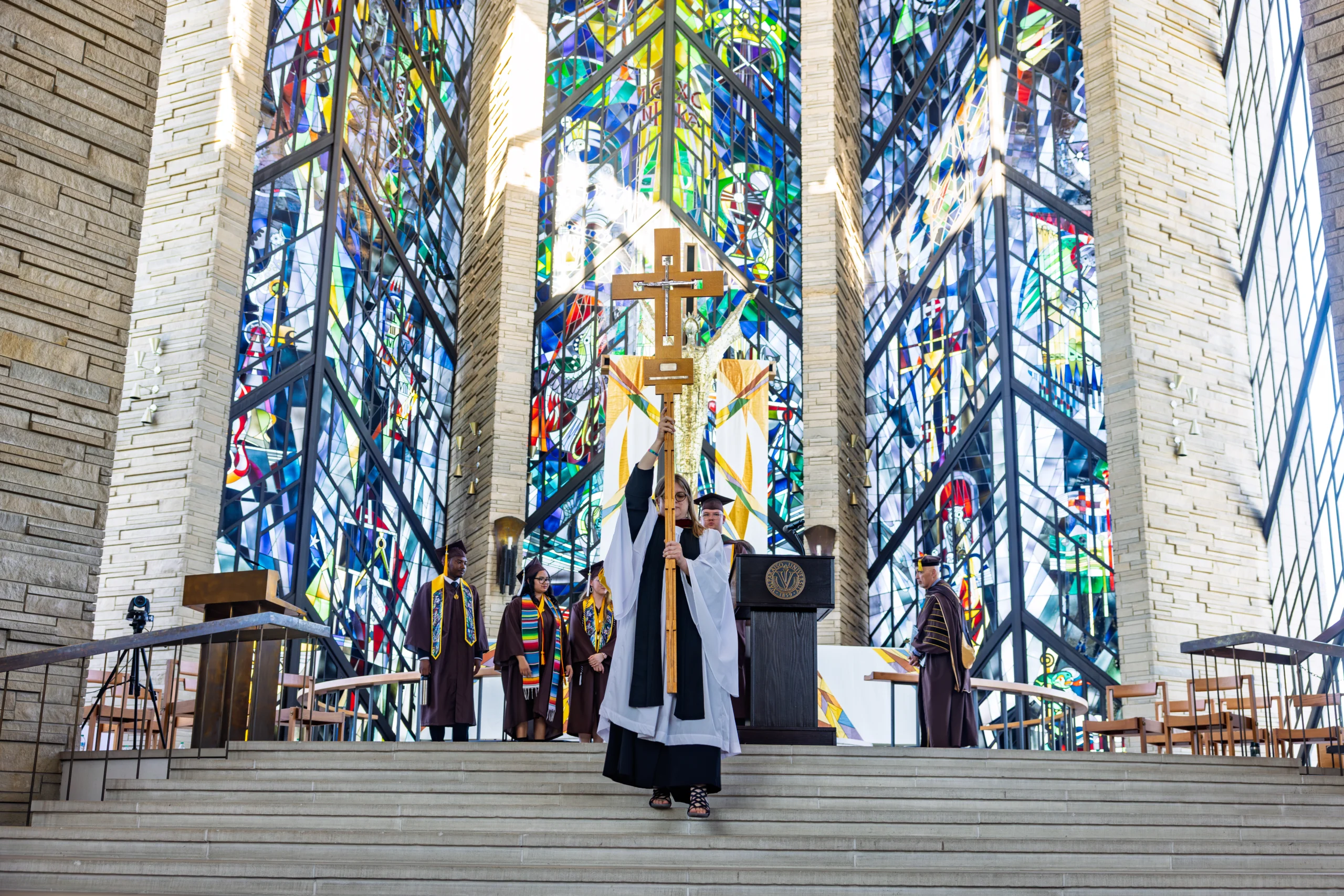 About: A person in clerical robes holds a cross in a church with colorful stained glass windows and graduates behind.
