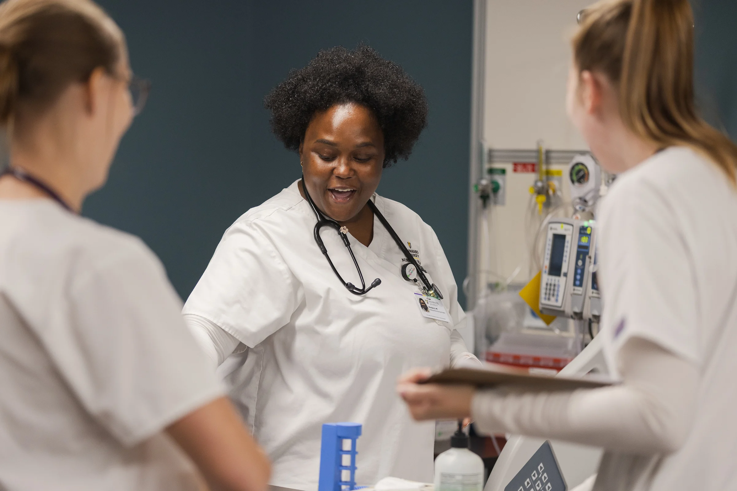 Three healthcare workers in white uniforms, one holding a clipboard, talk together in a hospital room—showcasing the teamwork valued by the College of Nursing and Health Professions.