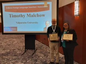 Two people holding certificates stand by a screen displaying “2025 Teacher of the Year: Timothy Malchow,” celebrating Valparaiso University’s tradition of excellence in German Education.