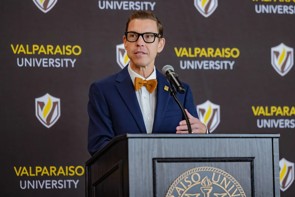 A man in a bow tie speaks at a podium with "Valparaiso University" and its logo in the background, marking an exciting moment for Valpo as the new president addresses campus life and future initiatives.