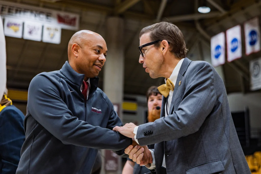 Two men shake hands and talk at a Valpo basketball game, one in a suit and bow tie—perhaps the New President—while the other is in athletic wear, capturing the spirit of campus life.