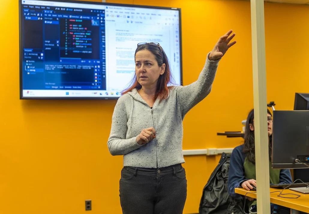 A woman gestures while teaching in a classroom at Valparaiso University, with a coding screen displayed on a large monitor behind her, highlighting the AI Practitioner Certificate program.