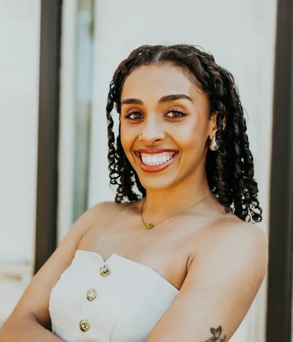 Smiling woman, Brielle Wilkins, with curly hair and a floral tattoo poses confidently in a white strapless dress.