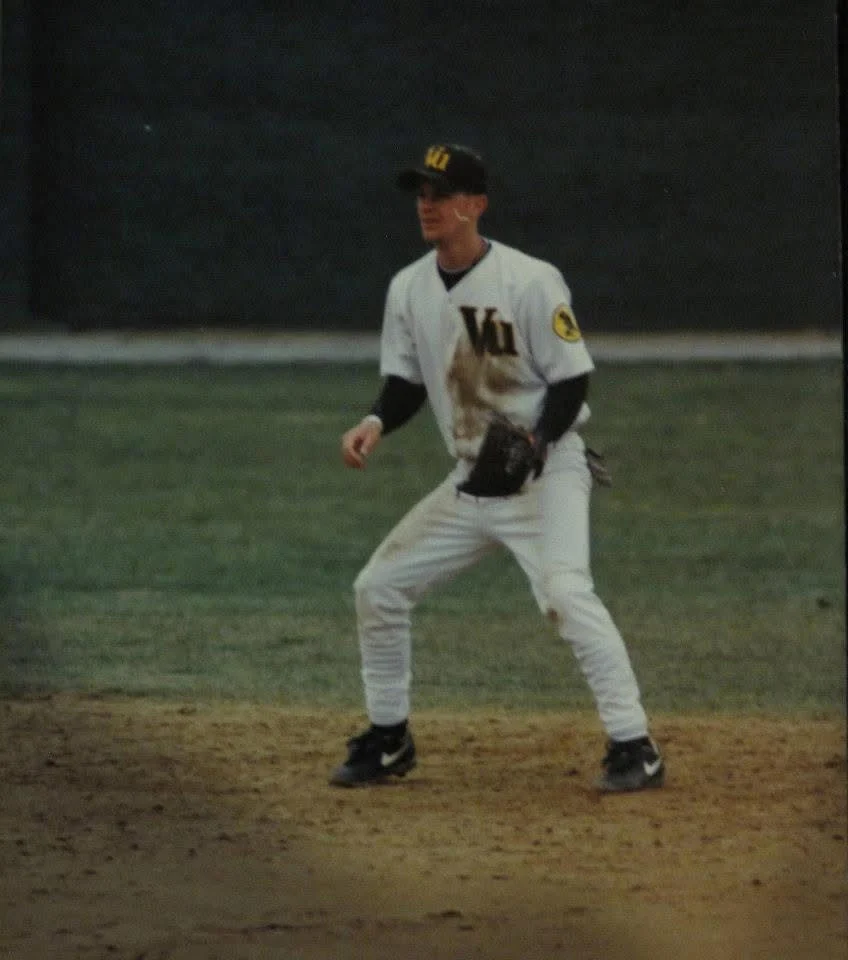 Baseball player Jeff Conrad in a white uniform fields on a dirt infield, ready in an athletic stance, acting as a beacon of focus and determination.