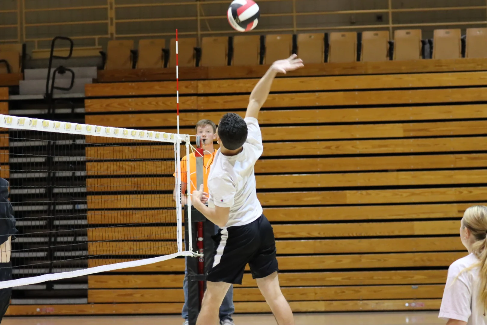 A volleyball player jumps to spike the ball during intramurals in an indoor gym, with a referee watching closely.