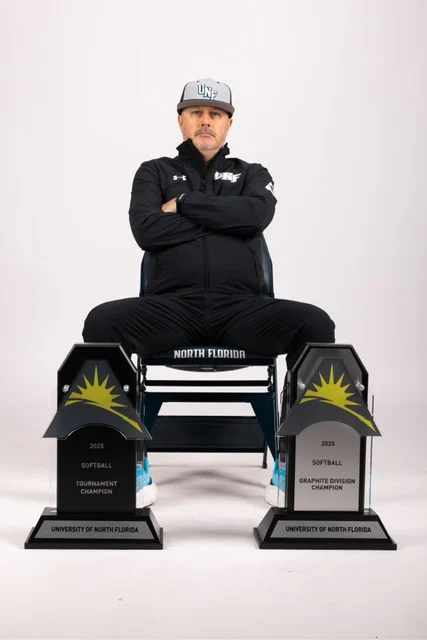 Jeff Conrad, dressed in a UNF tracksuit and cap, sits proudly with two Beacon softball championship trophies at his feet.