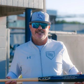 Jeff Conrad, sporting sunglasses and UNF baseball gear, stands by the dugout with a bat in hand, smiling like a true Beacon of team spirit.