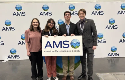 Three Valparaiso University meteorology students and a professor stand together, smiling while holding an American Meteorological Society Annual Meeting sign.