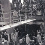 Crowds of immigrants gather on a ship’s deck, some standing by railings, others sitting with belongings.