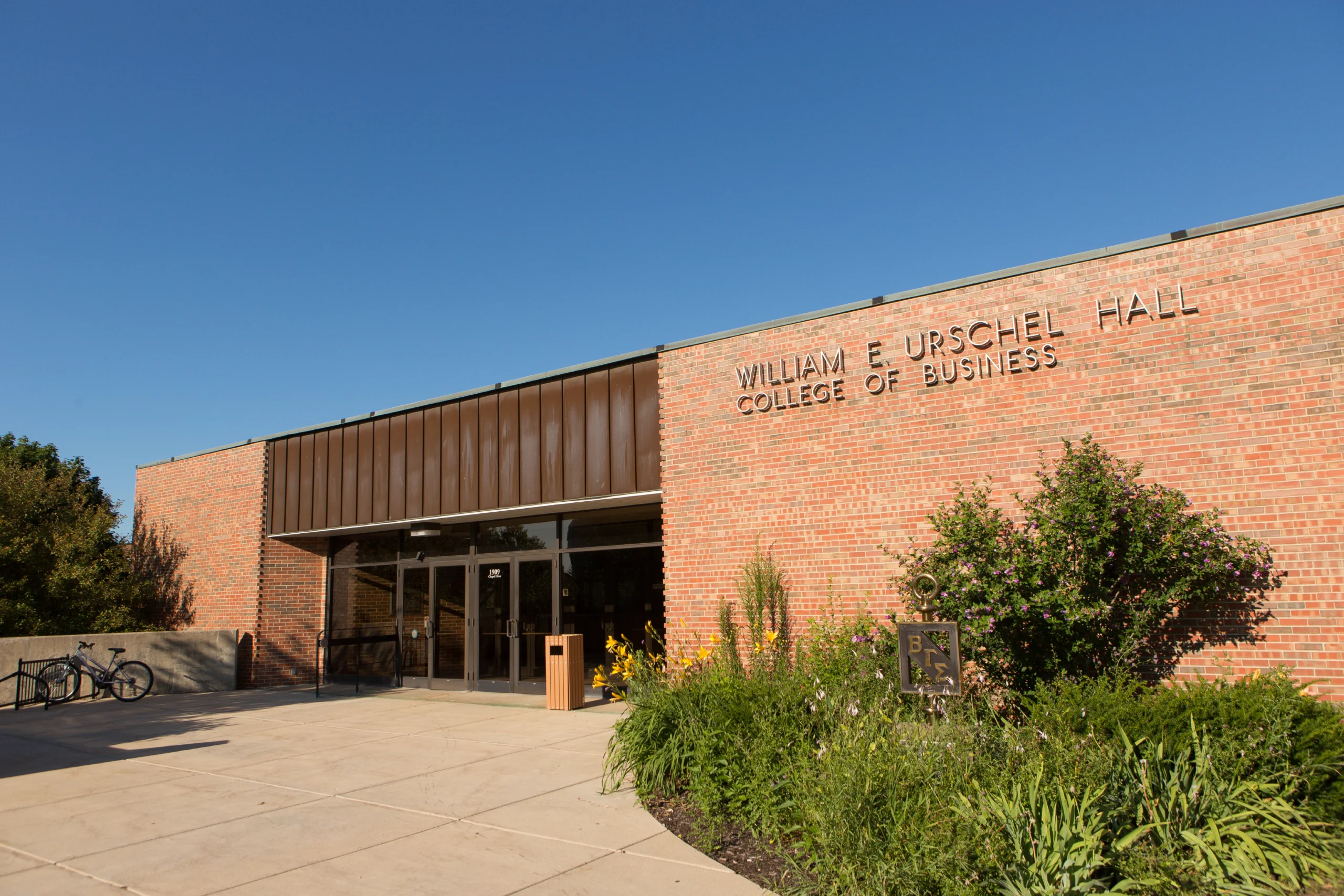 Brick building entrance with “William E. Urschel Hall College of Business” sign, plants, and clear sky—where the spirit of innovation shines like Beacon’s Reflection. Looking ahead to 2027, students like Esai Hernandez will continue to shape the future within these inspiring walls.