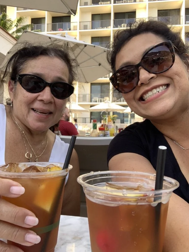 Two women smiling and holding drinks outdoors, sitting at a table near a hotel pool area with umbrellas, enjoy a conversation about Izzy Ibarra’s MBA journey from Beacon's Reflection.