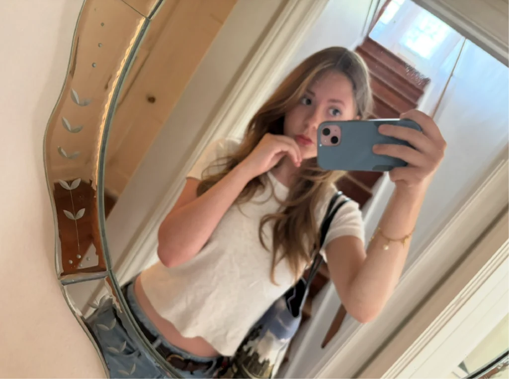 Young woman takes a mirror selfie with her phone, wearing a white crop top and carrying a bag, capturing Beacon's Reflection in the background.