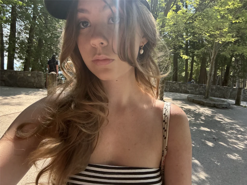 Young woman in a striped top and black cap takes a selfie outdoors in a sunny, tree-filled park, capturing the spirit of Beacon's Reflection.