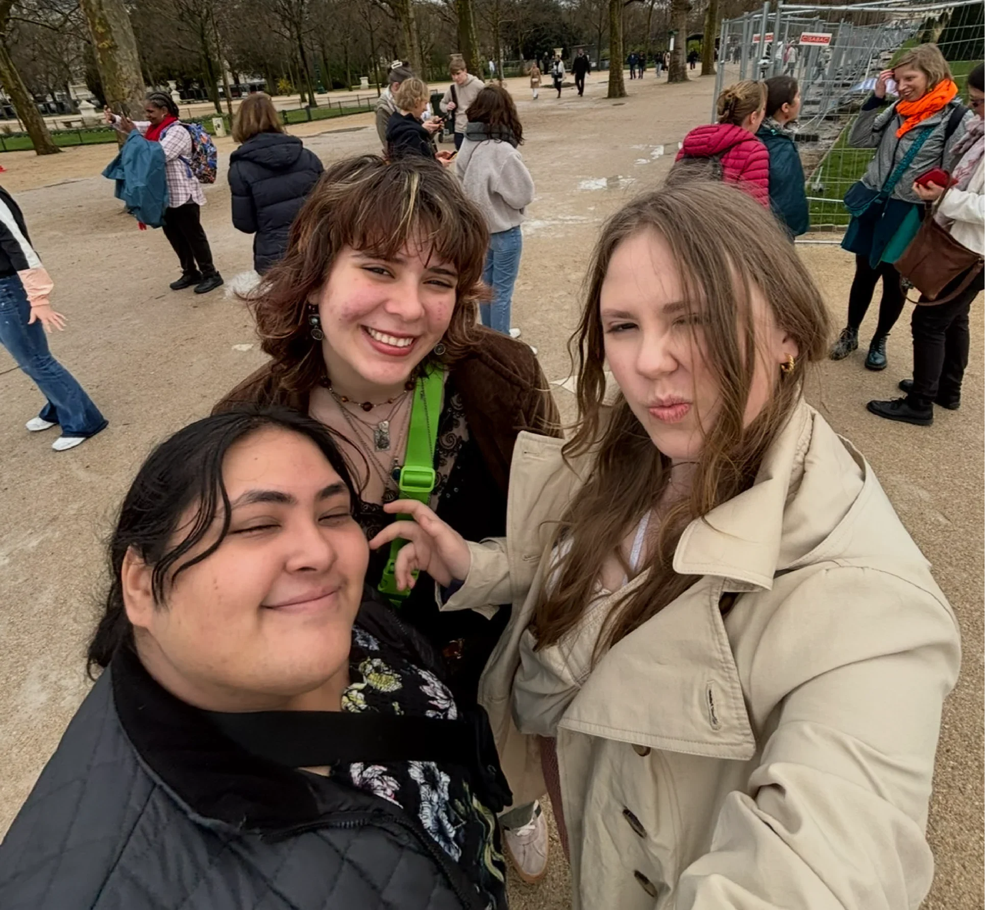 Three smiling friends take a selfie near the Eiffel Tower on a cloudy day, capturing Bryn Solmos’s adventurous spirit amid trees and tourists—a true Beacon's Reflection of friendship in Paris.