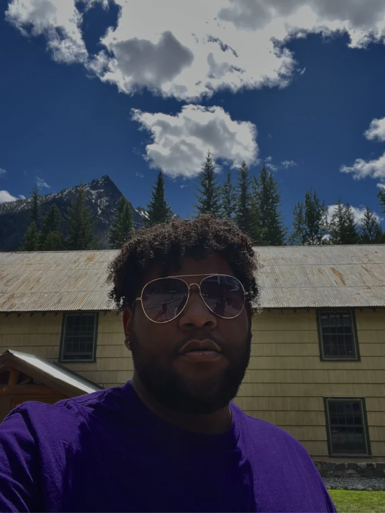 Esai Hernandez, in sunglasses and a purple shirt, stands outdoors with a rustic building, trees, mountains, and blue sky behind—a scene reminiscent of Beacon's Reflection.