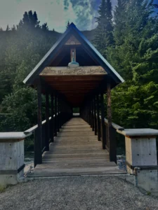 A wooden covered bridge in the forest, with a sign reading "Where the river flows, life abounds," reflects the quiet wisdom of Esai Hernandez in Beacon's Reflection.