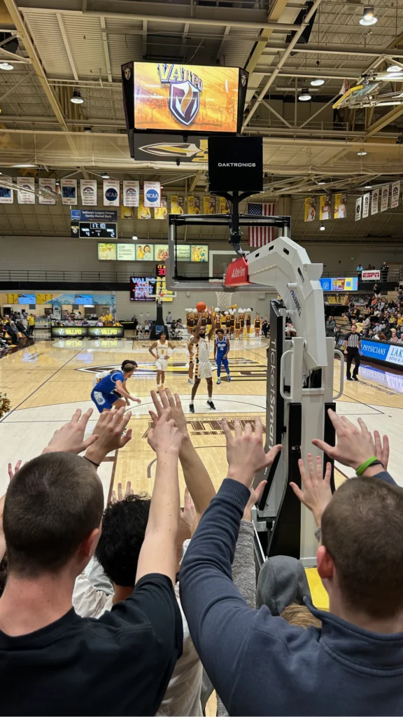 Fans raise their arms as Parker Kelson shoots a free throw in a college gym, with the mascot nearby and Beacon's Reflection shining from the scoreboard.