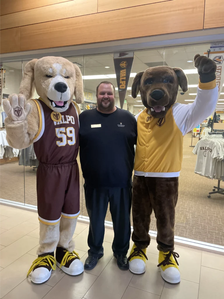 Parker Kelson stands between two people in dog mascot costumes, all smiling inside a campus store—capturing a moment of unity in Beacon's Reflection.