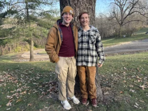 Two people stand by a tree on a leafy lawn, smiling, dressed in casual fall clothing—a scene reminiscent of Beacon’s Reflection. One of them could be Parker Kelson, enjoying the crisp autumn air with a friend.