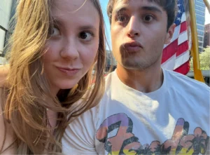 Two young adults take a selfie outdoors with an American flag visible in the background, capturing a moment that echoes Beacon's Reflection.