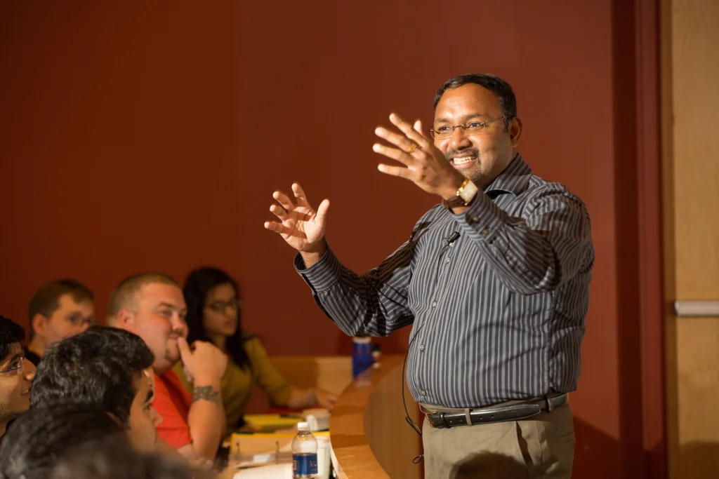 A man giving a lecture to a group of people in a classroom setting, gesturing with his hands as he explains the benefits of experiential learning.