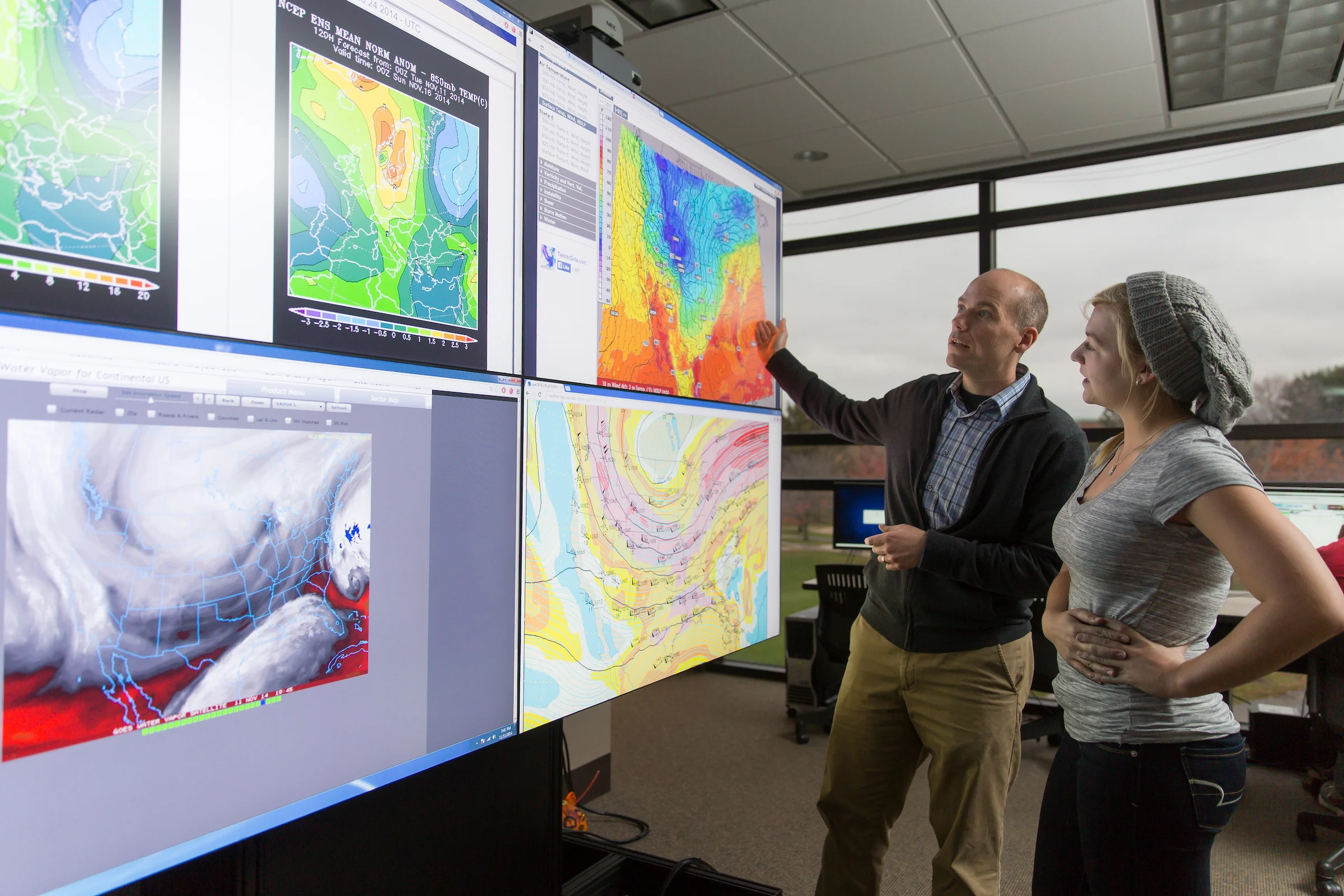 Two people analyze colorful weather maps on large computer screens in a modern meteorology office setting.