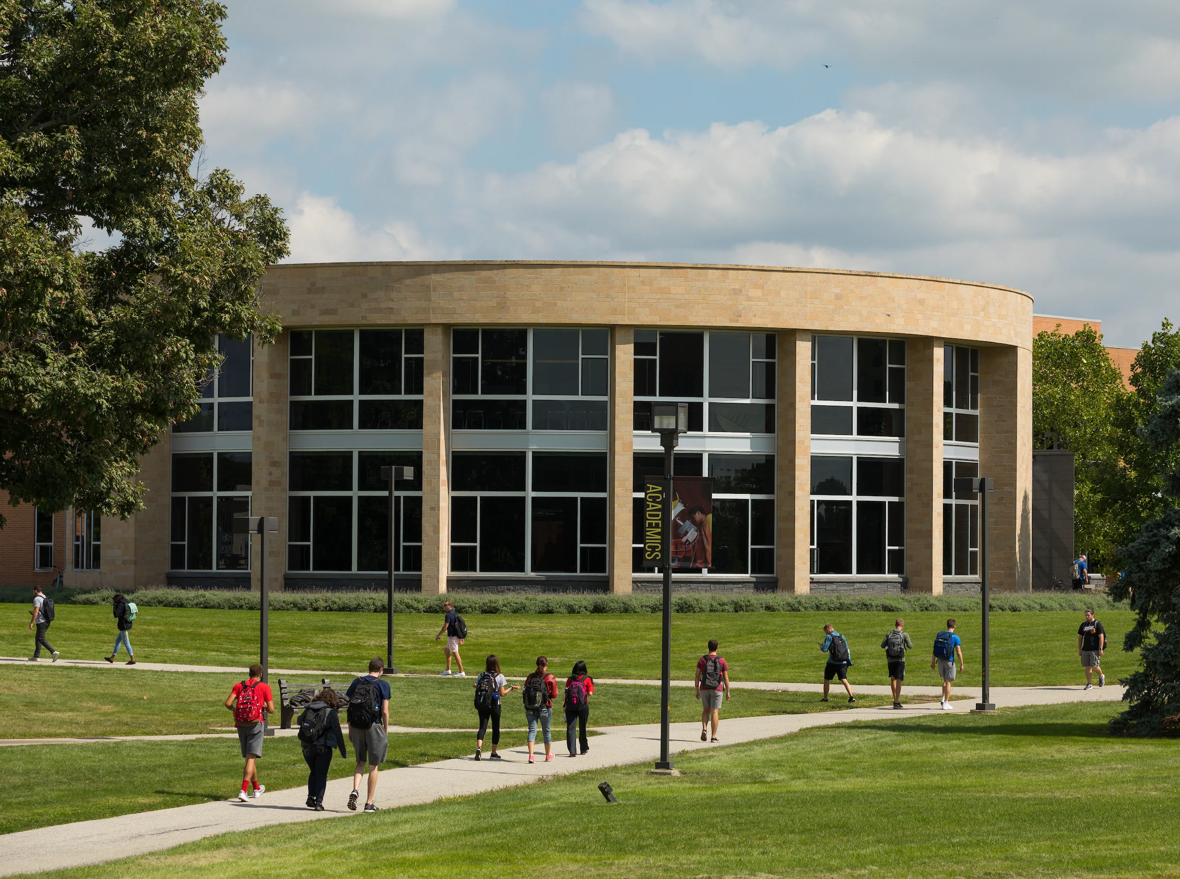 Students experience college life as they walk on paths across a grassy campus in front of a modern, round building with large windows at Valparaiso University.