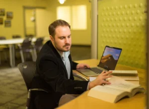 A man sitting at a desk with books and a laptop, reaching to turn a page in an open book—an inspiring scene of adult education and going back after 30 years.