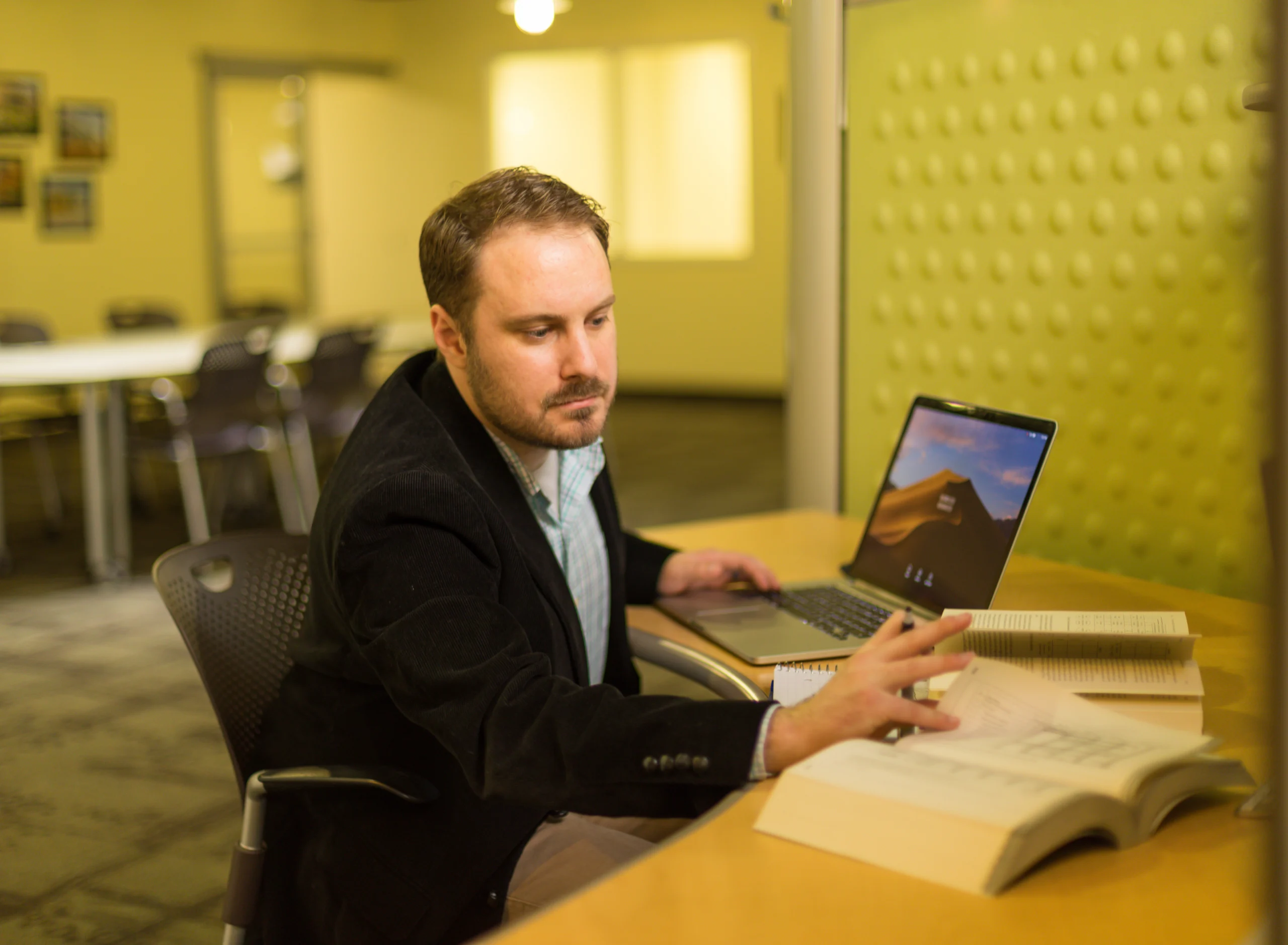 A man sitting at a desk with books and a laptop, reaching to turn a page in an open book—an inspiring scene of adult education and going back after 30 years.
