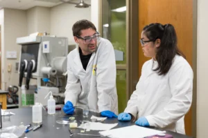 Two scientists in lab coats and gloves discuss an experiment at a laboratory bench with equipment and notes, reflecting the hands-on college experience shaped by a low student-to-faculty ratio.