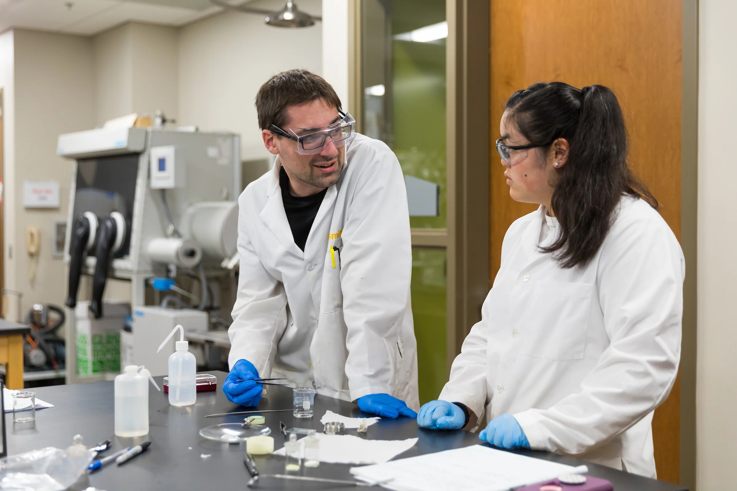Two scientists in lab coats and gloves discuss an experiment at a laboratory bench with equipment and notes, reflecting the hands-on college experience shaped by a low student-to-faculty ratio.