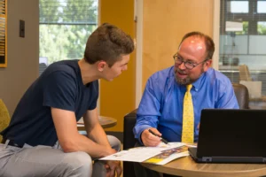 A man in a blue shirt and yellow tie talks with a young man over papers at a desk with a laptop.