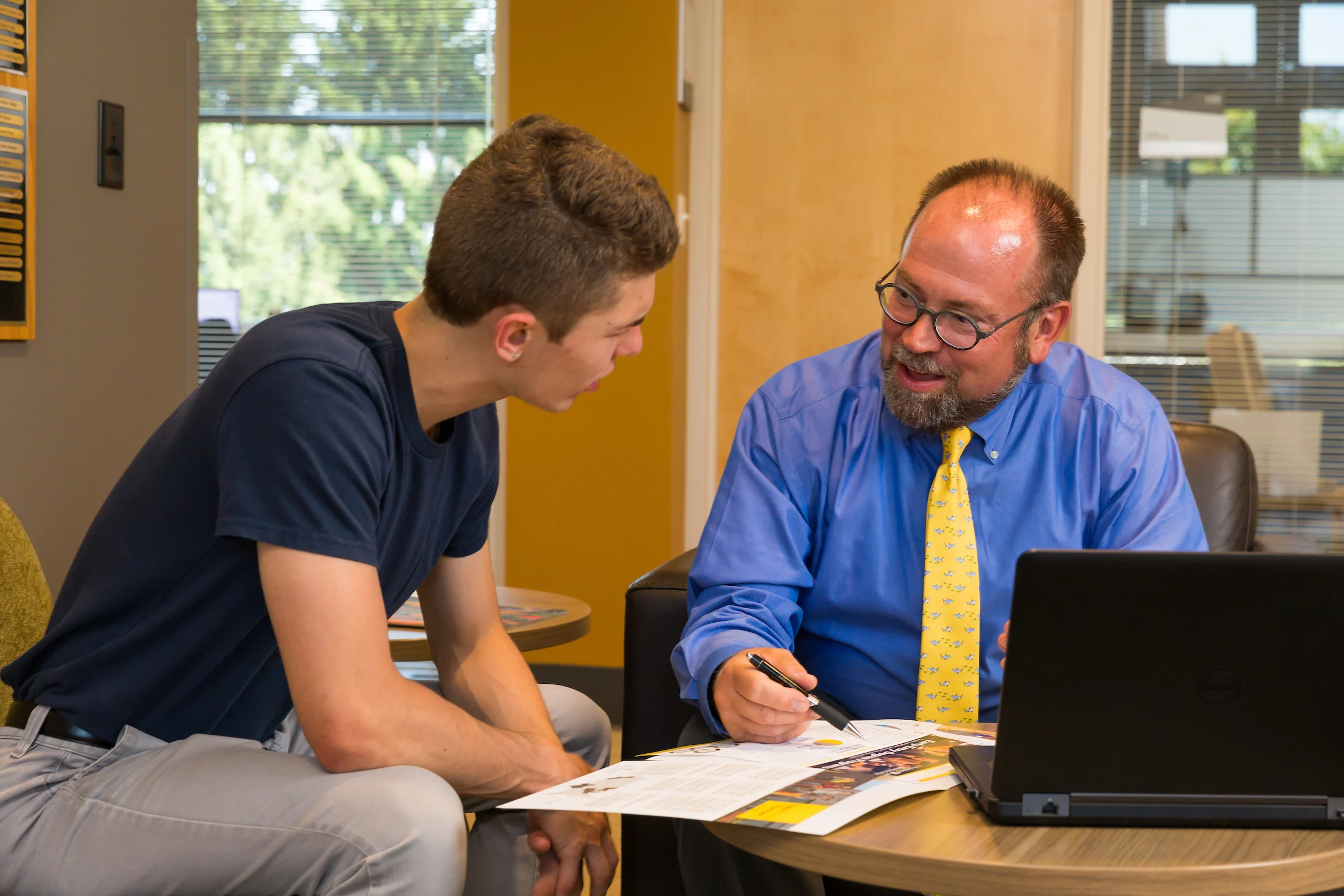 A man in a blue shirt and yellow tie talks with a young man over papers at a desk with a laptop.