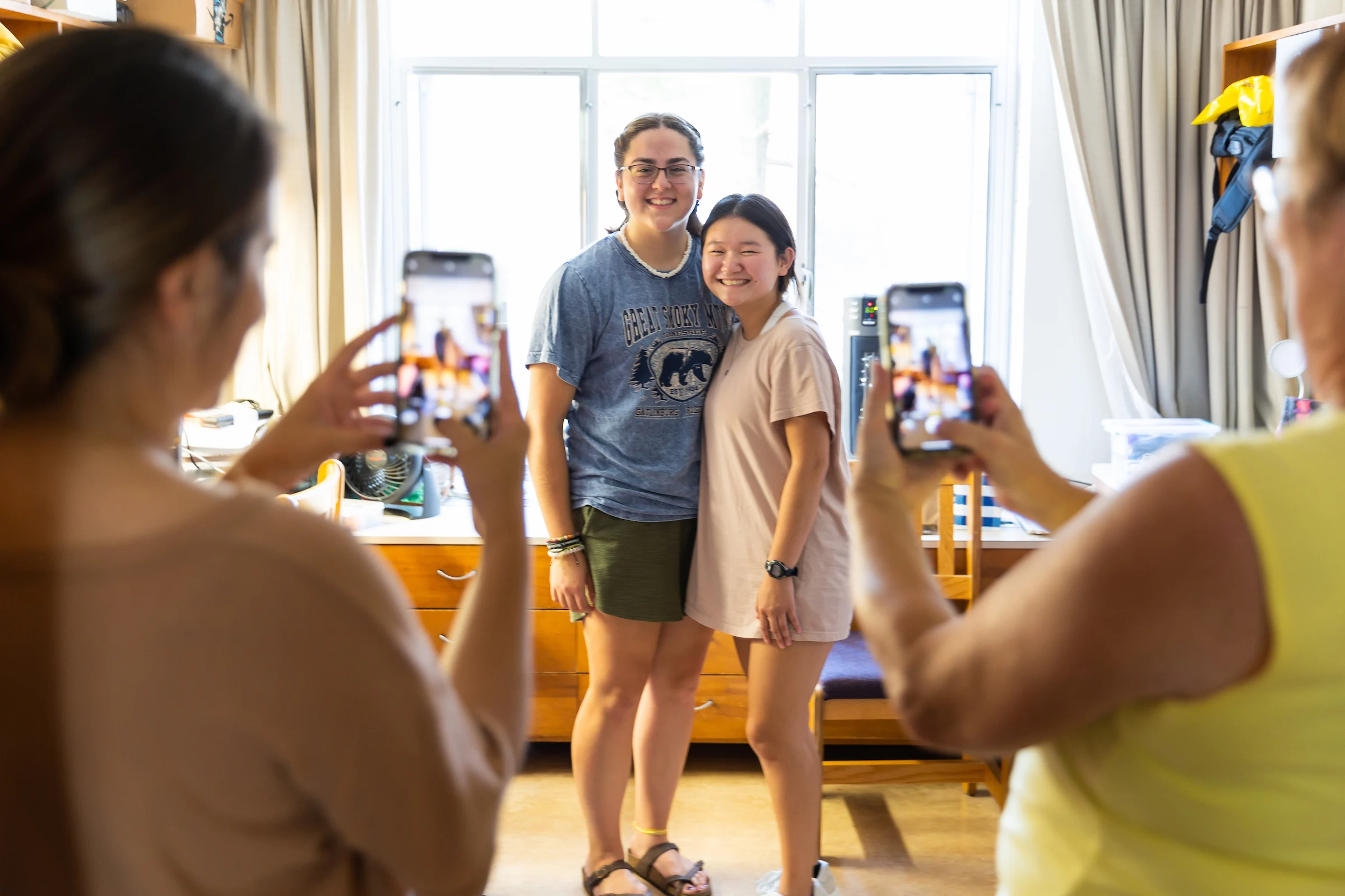 Two young women smile and pose for photos inside a bright room as others take pictures with smartphones.
