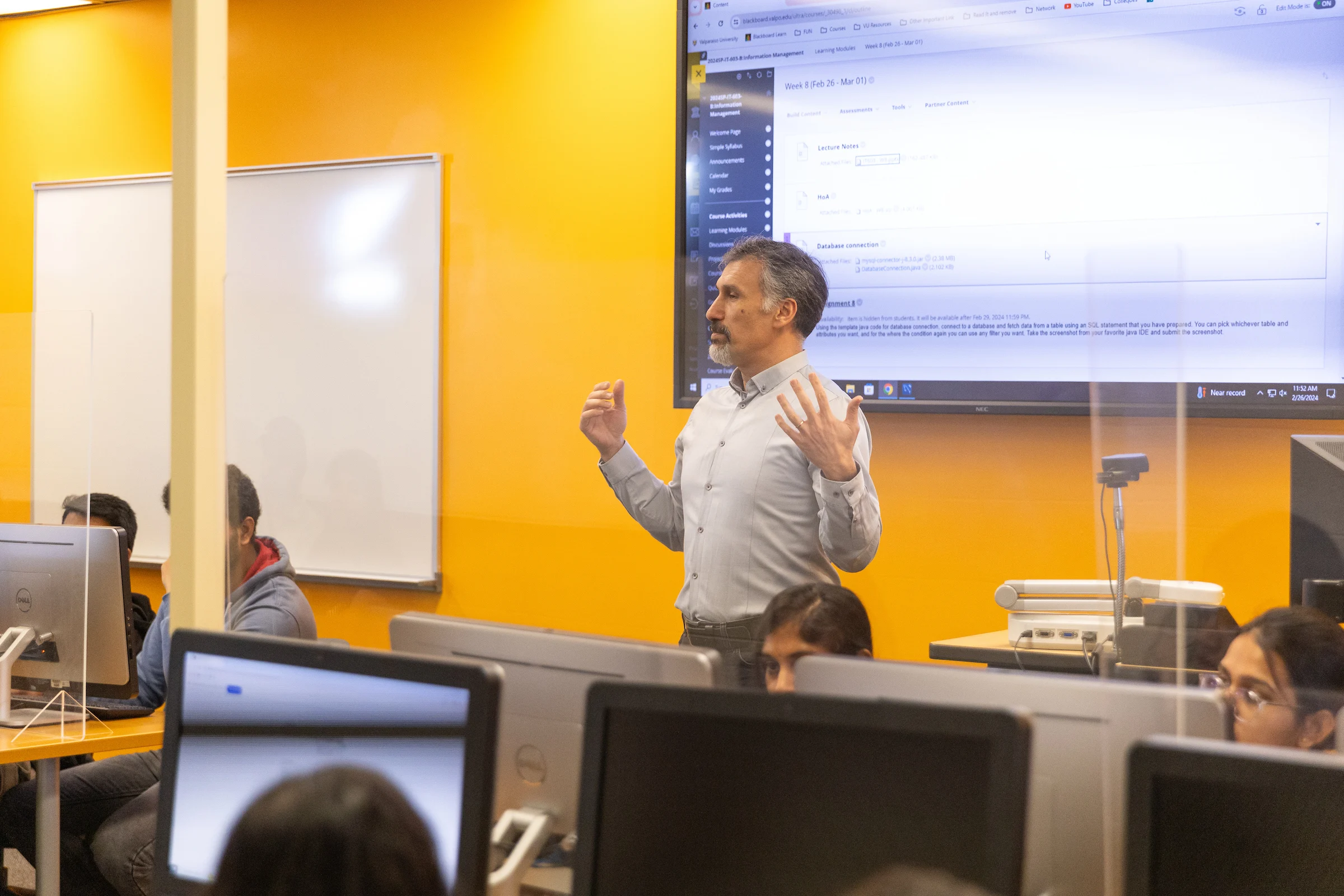 A man teaches a computer class at Valparaiso University, standing in front of monitors and a large screen in a bright classroom focused on the AI Practitioner Certificate.