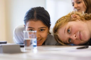 Two girls smiling and leaning on a table with a clear plastic cup of water in the foreground, capturing the excitement of what to expect during your freshman year of college.