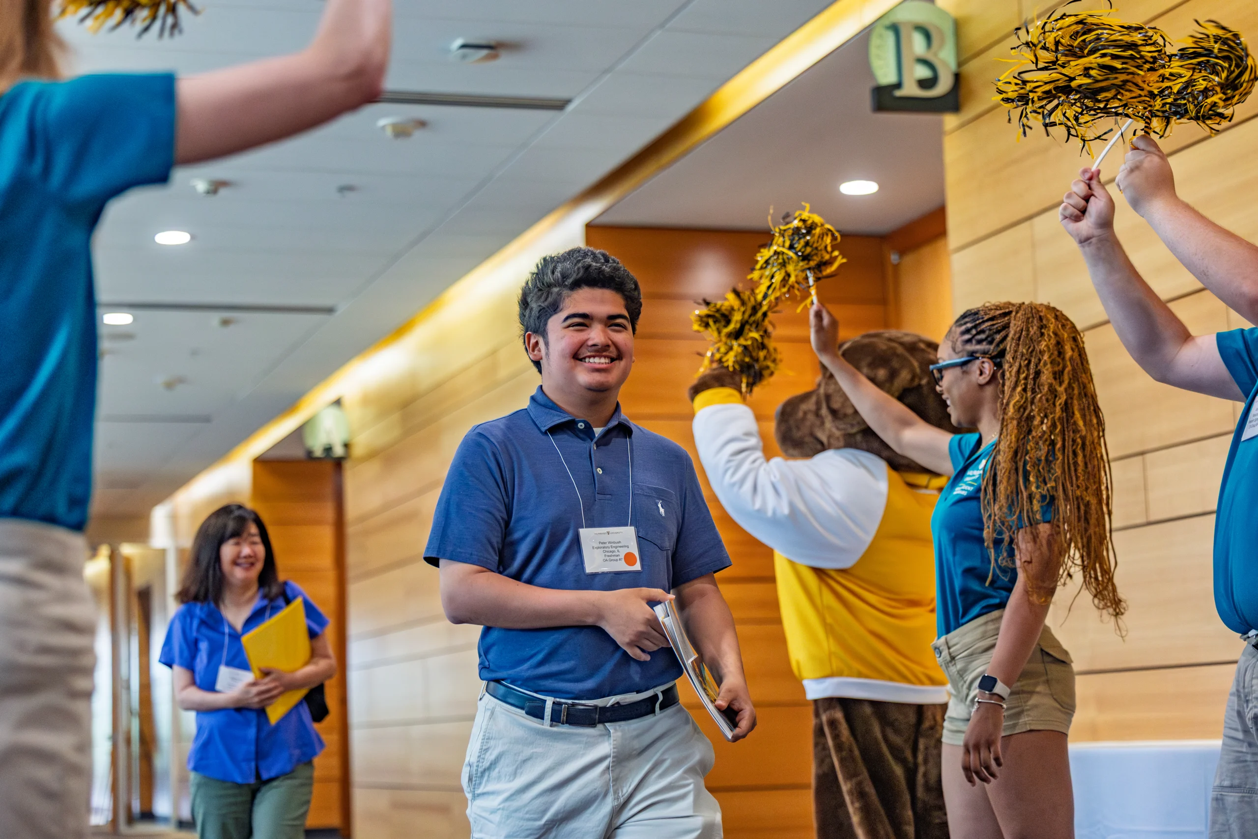 Smiling student walks down hallway as people cheer with yellow pom-poms, keeping their FOCUS on celebrating; a mascot is in the background.
