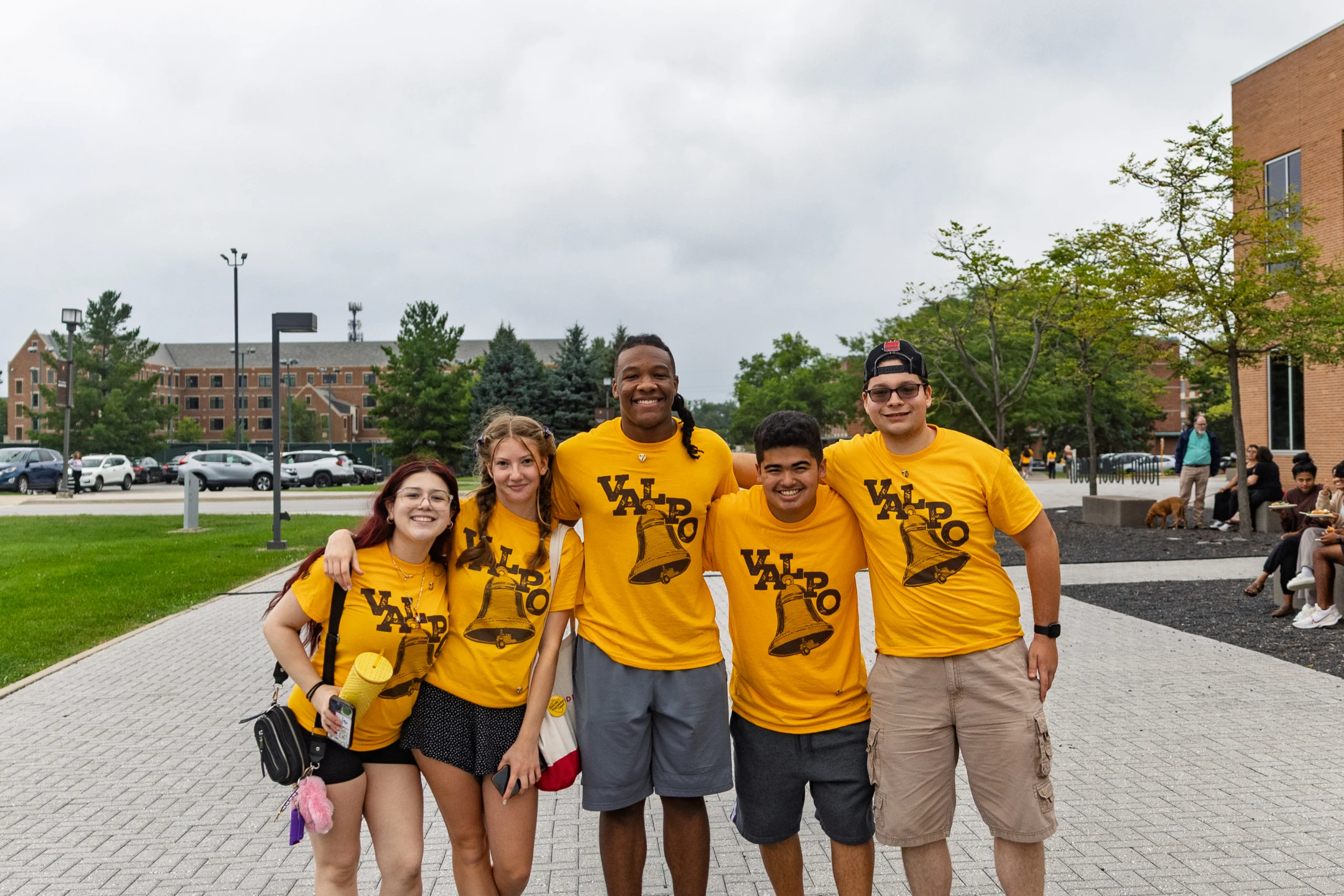 Five smiling students in matching yellow Valpo shirts pose together outside on a cloudy day, ready to welcome Parents and Families to campus.