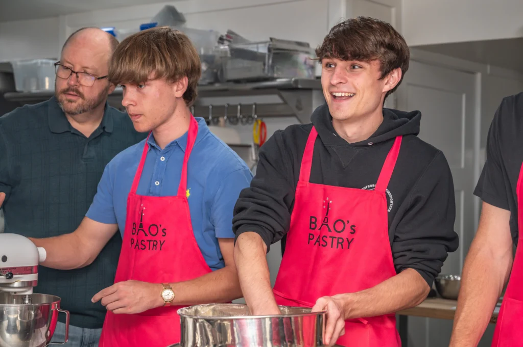 Three people wearing red aprons enjoy experiential learning while baking together in a kitchen, one smiling as they mix dough in a bowl—capturing the collaborative spirit of Valpo’s French program.