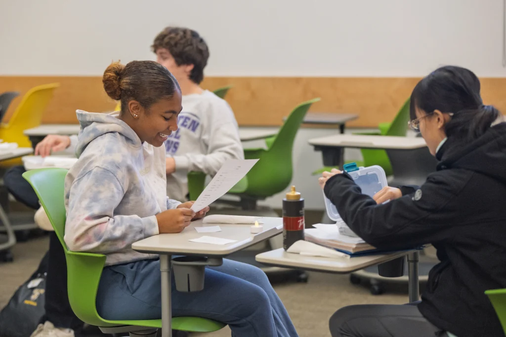 Two students sit at desks, smiling and reading papers in a Valpo classroom with green chairs, enjoying experiential learning in the French program.