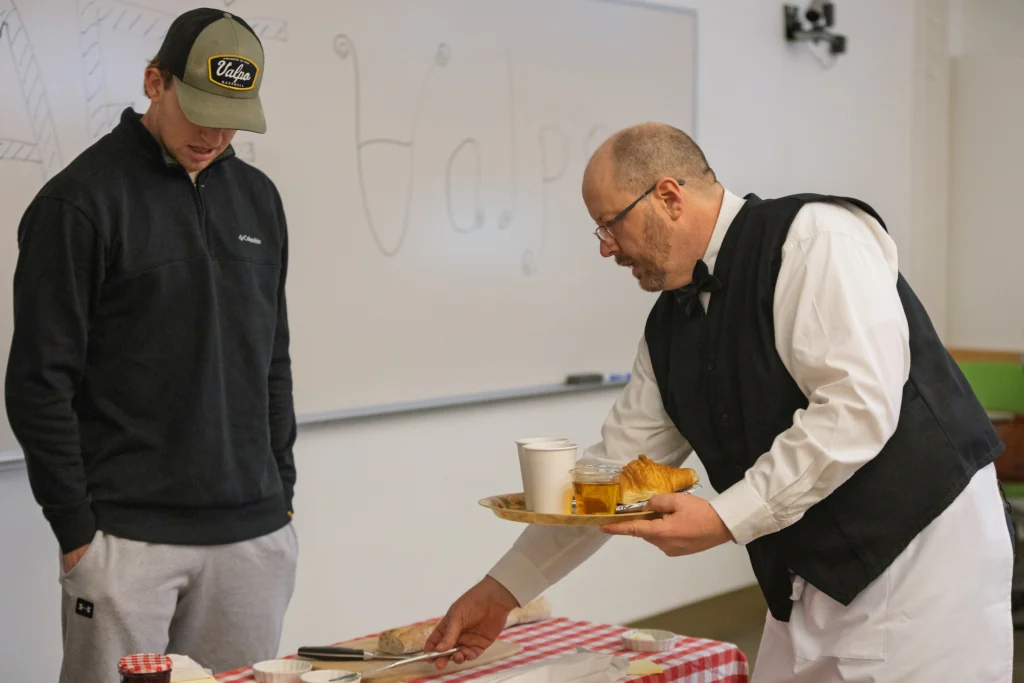 A man in a vest serves food to a person in casual clothes at a table with a red checkered tablecloth, showcasing the Valpo French program’s focus on experiential learning.