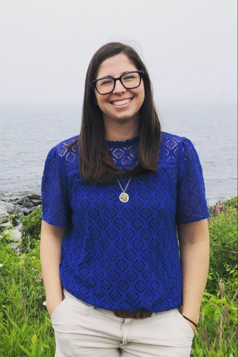 Woman with long brown hair, glasses, and a blue top smiles outdoors near greenery and a body of water, perhaps enjoying one of the local workshops held in this scenic setting.