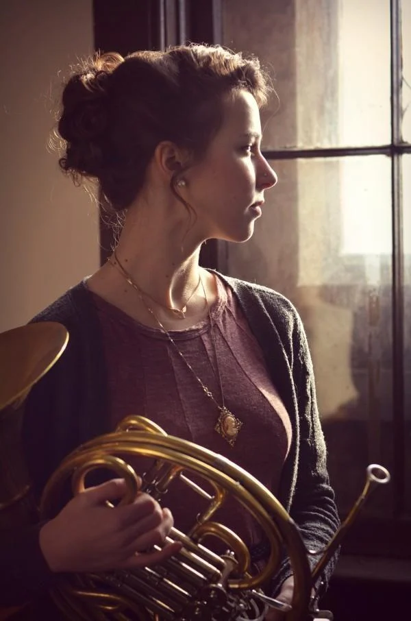 A woman holding a French horn stands by a window, looking thoughtfully into the distance, perhaps reflecting on recent workshops she attended.