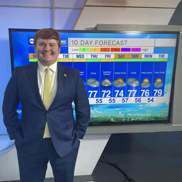 Smiling man in a suit, likely Emmett Twomey, stands by a TV displaying a 10-day weather forecast in a news studio, showcasing his passion for meteorology.