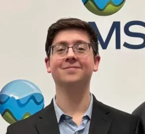 Ethan Palmisano, in glasses and a suit, stands in front of a white background with a blue and green logo.