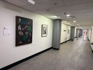 A hallway art gallery at Christopher Center Library, Valparaiso University, features framed artwork hanging on white walls and gray carpeted floors for the Stories Art Exhibition.