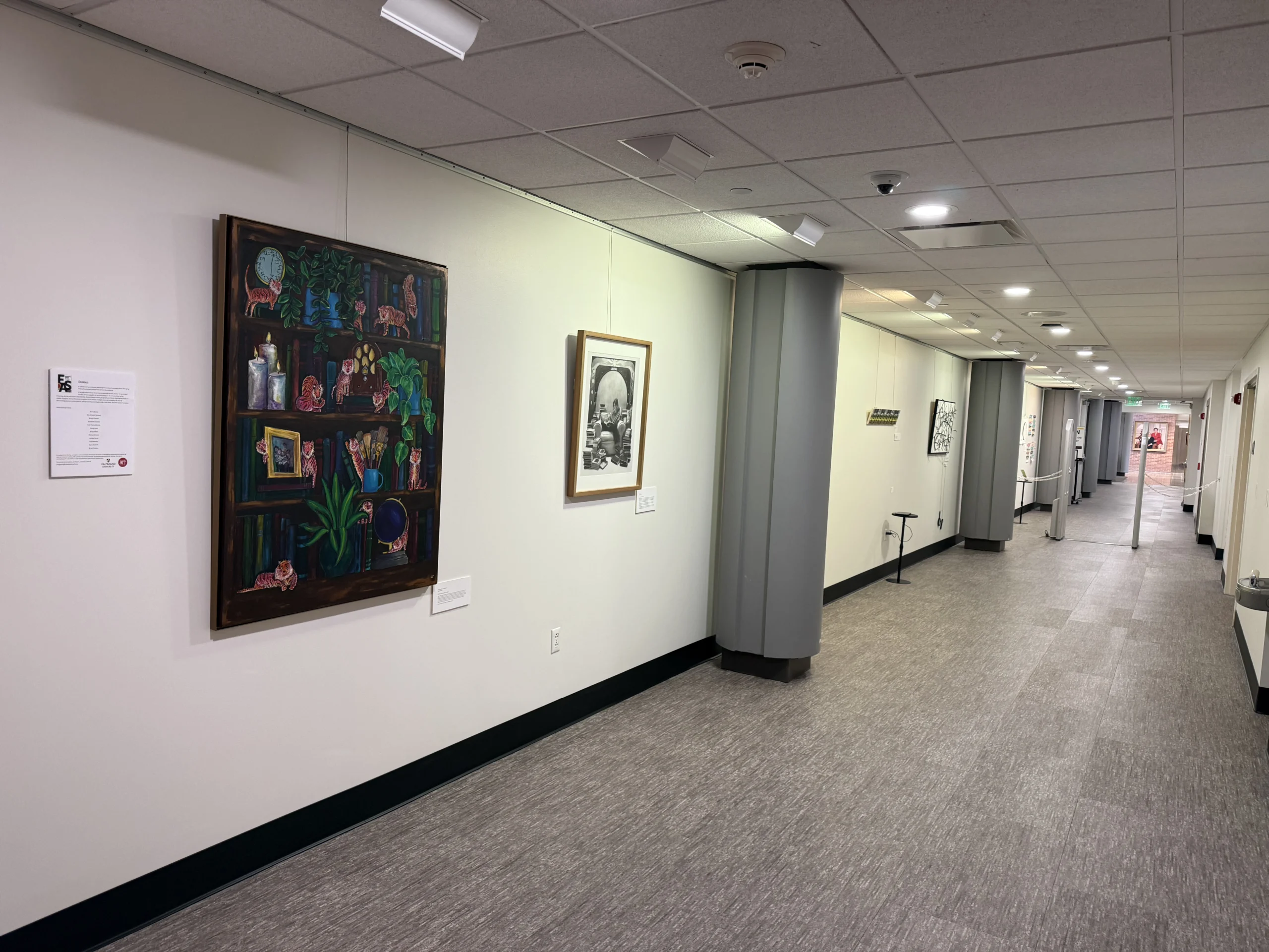 A hallway art gallery at Christopher Center Library, Valparaiso University, features framed artwork hanging on white walls and gray carpeted floors for the Stories Art Exhibition.
