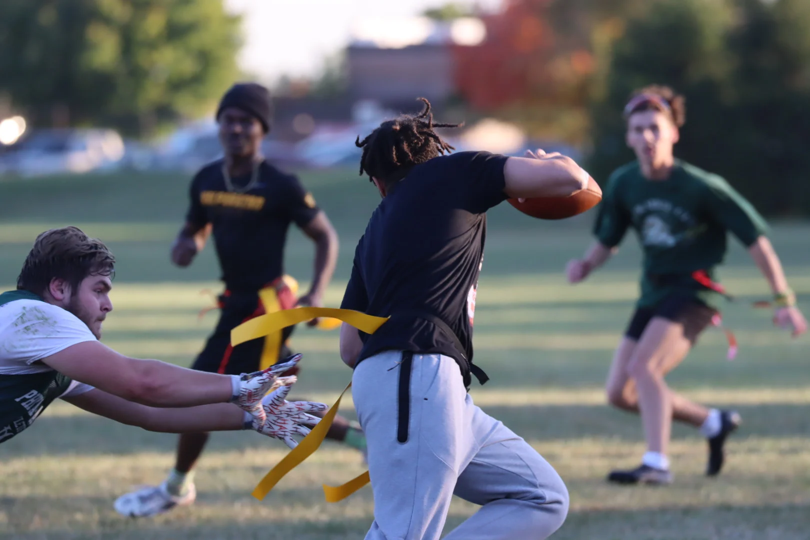 An intramurals flag football player runs with the ball as a defender tries to grab his flag on a grassy field.